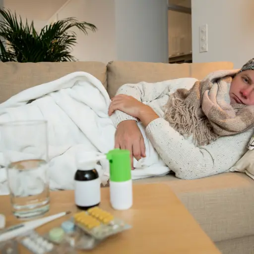 Sick man wearing scarf lying on couch at home under a blanket checking temperature. Living room with table full of medicine and pills. Man with running nose and cough.